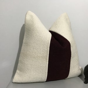 May include: Cream-colored decorative pillow with a vertical burgundy stripe. The pillow has a textured weave and a square shape, perfect for adding a touch of color and texture to any room. A small, dark elephant figurine is in the background.