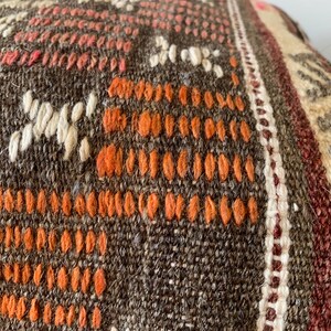 May include: Close-up of a woven textile with a brown, orange, and cream color palette. The fabric features a detailed pattern of geometric shapes and lines, with a textured surface. The design includes orange, white, and pink accents.