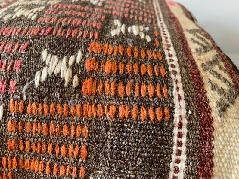 May include: Close-up of a woven textile with a brown, orange, and cream color palette. The fabric features a detailed pattern of geometric shapes and lines, with a textured surface. The design includes orange, white, and pink accents.