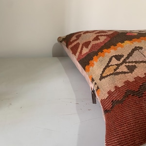 May include: A colourful throw pillow with a geometric pattern in shades of red, orange, brown, and beige. The pillow is made of woven fabric and has a fringed edge.