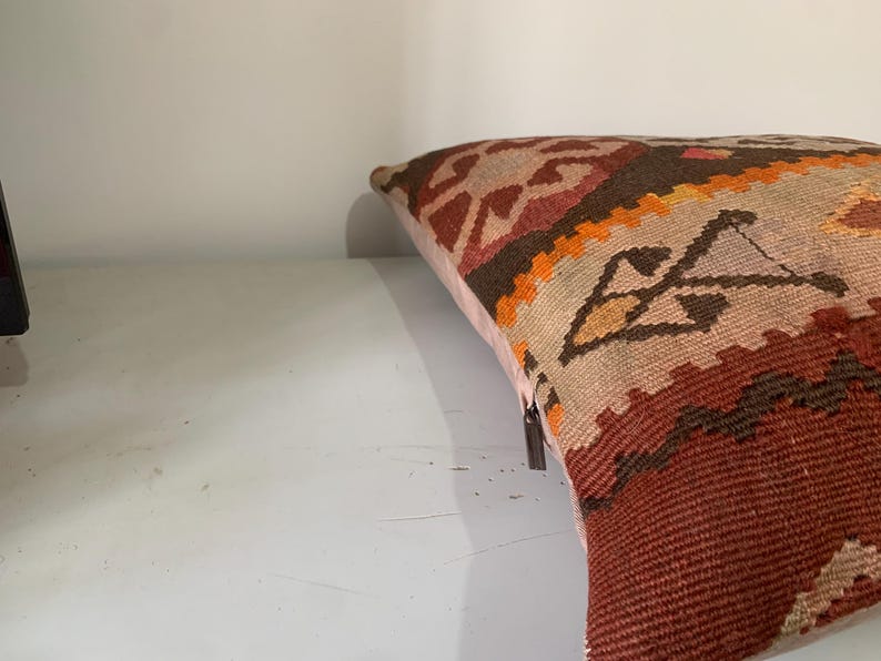 May include: A colourful throw pillow with a geometric pattern in shades of red, orange, brown, and beige. The pillow is made of woven fabric and has a fringed edge.