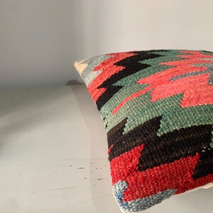 May include: A colorful throw pillow with a geometric pattern in shades of red, green, black, and white. The pillow is made of woven fabric and has a fringed edge.
