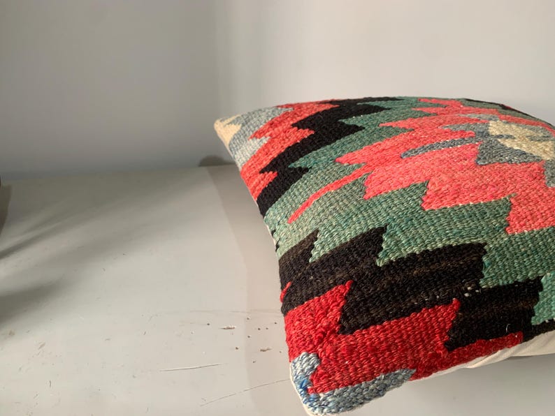 May include: A colorful throw pillow with a geometric pattern in shades of red, green, black, and white. The pillow is made of woven fabric and has a fringed edge.