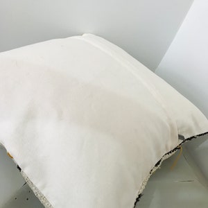 Puede incluir: Almohada blanca con ribete de flecos blanco y negro. La almohada est&aacute; sobre una superficie de madera.