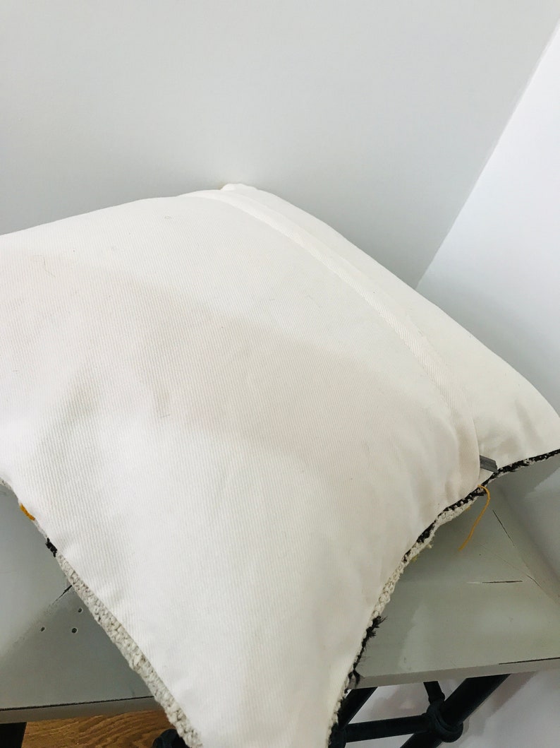 Puede incluir: Almohada blanca con ribete de flecos blanco y negro. La almohada est&aacute; sobre una superficie de madera.