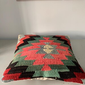May include: A colorful, geometric patterned pillow with red, green, black, and white colors. The pillow is made of woven fabric and has a traditional design.