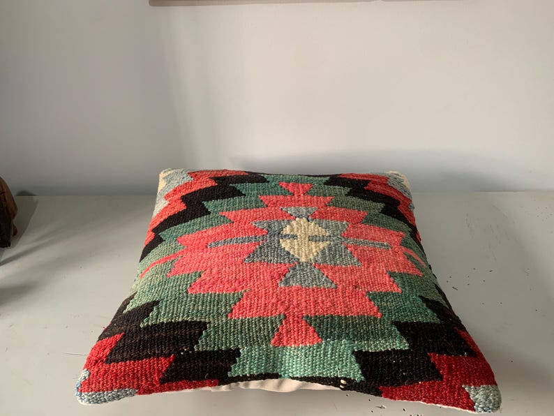 May include: A colorful, geometric patterned pillow with red, green, black, and white colors. The pillow is made of woven fabric and has a traditional design.