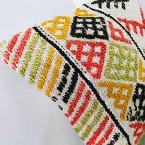 Puede incluir: Una almohada blanca con un patr&oacute;n geom&eacute;trico colorido en tonos de rojo, naranja, amarillo, verde y negro. El patr&oacute;n est&aacute; tejido en la tela y crea un aspecto texturizado.