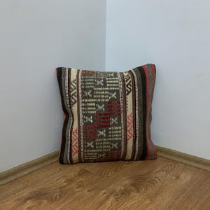 May include: Square decorative pillow with a woven geometric pattern in shades of brown, red, and cream. The pillow has a textured surface and is placed in a corner against a white wall and wooden floor.