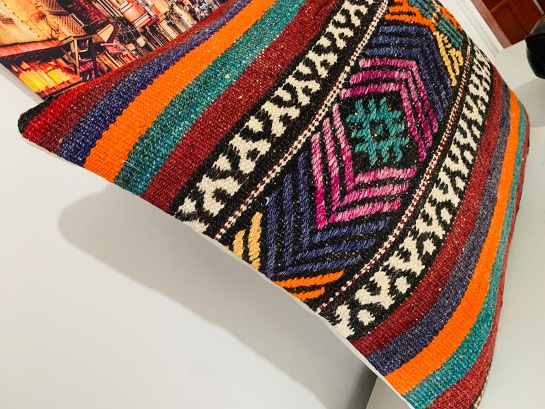 May include: A colorful throw pillow with a geometric pattern in shades of orange, red, blue, green, and white. The pillow is made of woven fabric and has a fringed edge.