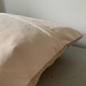 May include: Close-up of a beige pillow with a textured, woven border. The pillow has a soft, neutral color and a simple design, suitable for various home decor styles. The pillow is resting on a light gray surface.