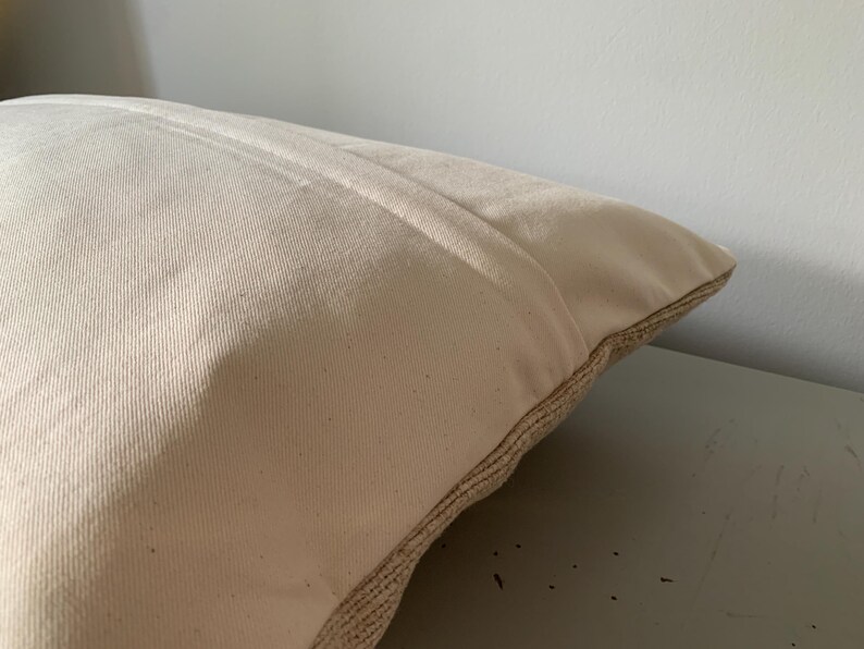 May include: Close-up of a beige pillow with a textured, woven border. The pillow has a soft, neutral color and a simple design, suitable for various home decor styles. The pillow is resting on a light gray surface.