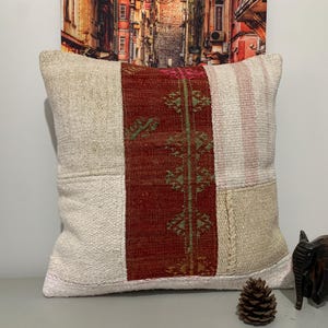 May include: A square decorative pillow with a patchwork design. The pillow features a central vertical panel of deep red fabric with green and gold embroidered details. The surrounding panels are beige and off-white.