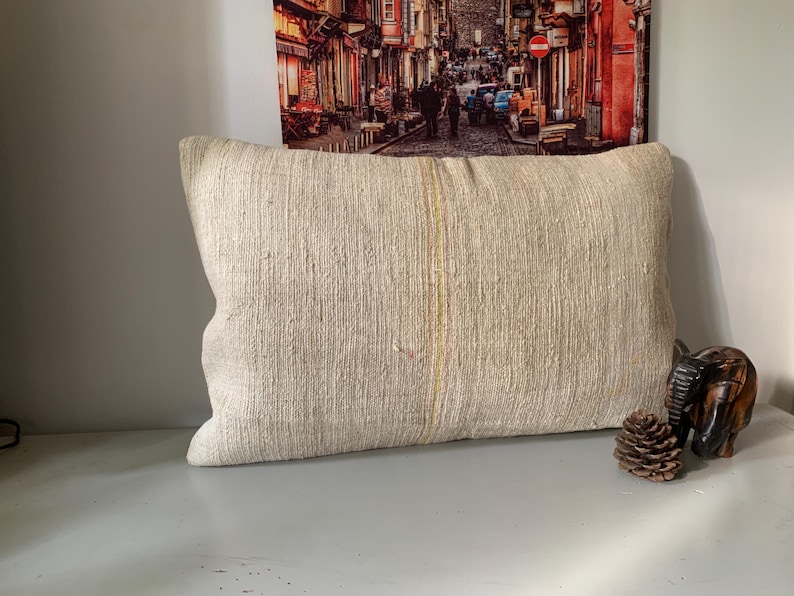 May include: Cream-colored rectangular pillow with a textured surface. The pillow has a vertical yellow stripe down the center. A wooden elephant figurine and a pine cone are next to the pillow.