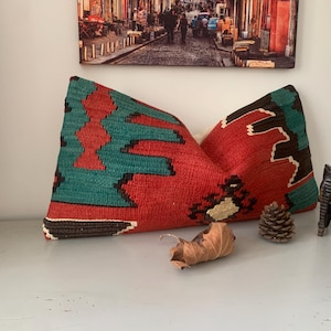 May include: A red, green, and black patterned throw pillow with a fringe border. The pillow is on a white surface with a small pine cone and a wooden elephant figurine.