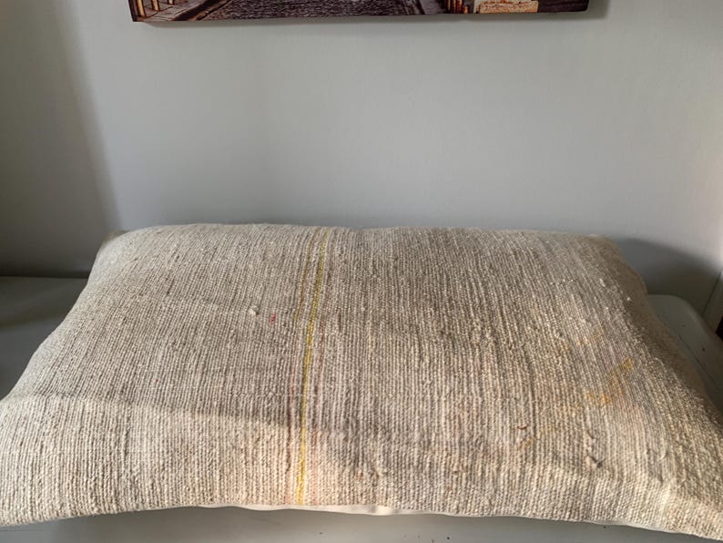 May include: A rectangular beige pillow with a textured, woven appearance. The pillow has a subtle vertical stripe of yellow and red. The pillow is a neutral color and would complement many decor styles.