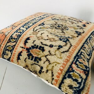 May include: A decorative pillow with a beige background and a colorful floral pattern in shades of blue, orange, and brown. The pillow has a white border.