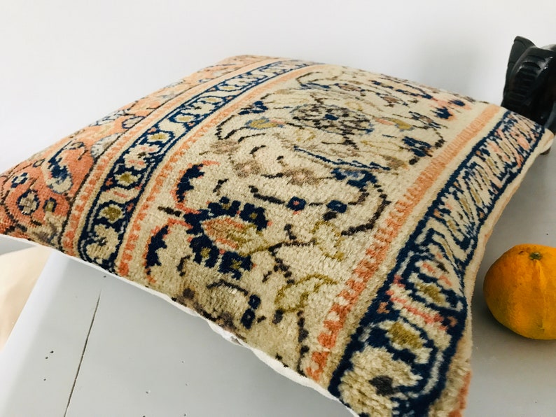 May include: A decorative pillow with a beige background and a colorful floral pattern in shades of blue, orange, and brown. The pillow has a white border.