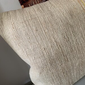 May include: Close-up of a beige woven pillow with a textured surface. The pillow has a rectangular shape and appears to be made of natural fibers. Thin yellow and red stripes run along one edge. The pillow is likely used for home decor.