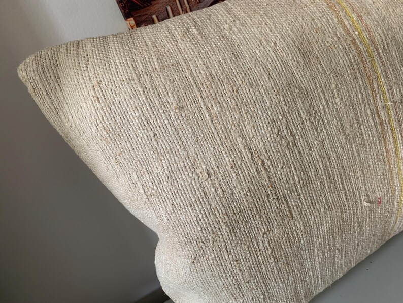 May include: Close-up of a beige woven pillow with a textured surface. The pillow has a rectangular shape and appears to be made of natural fibers. Thin yellow and red stripes run along one edge. The pillow is likely used for home decor.