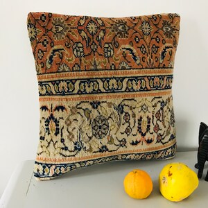 May include: A decorative pillow with a floral pattern in shades of beige, brown, and pink. The pillow has a border of dark blue and beige. The pillow is sitting on a white surface with a wooden elephant figurine and two yellow fruits.