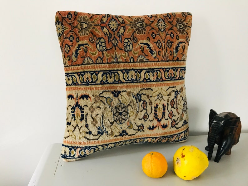 May include: A decorative pillow with a floral pattern in shades of beige, brown, and pink. The pillow has a border of dark blue and beige. The pillow is sitting on a white surface with a wooden elephant figurine and two yellow fruits.