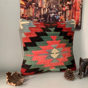 May include: A colorful throw pillow with a geometric pattern in shades of red, green, black, and white. The pillow is made of woven fabric and has a fringed edge.