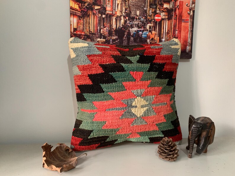 May include: A colorful throw pillow with a geometric pattern in shades of red, green, black, and white. The pillow is made of woven fabric and has a fringed edge.