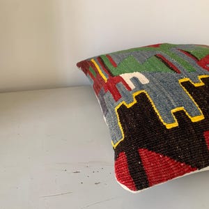 May include: A colorful throw pillow with a geometric pattern in shades of red, green, blue, black, and yellow. The pillow is made of woven fabric and has a white border.