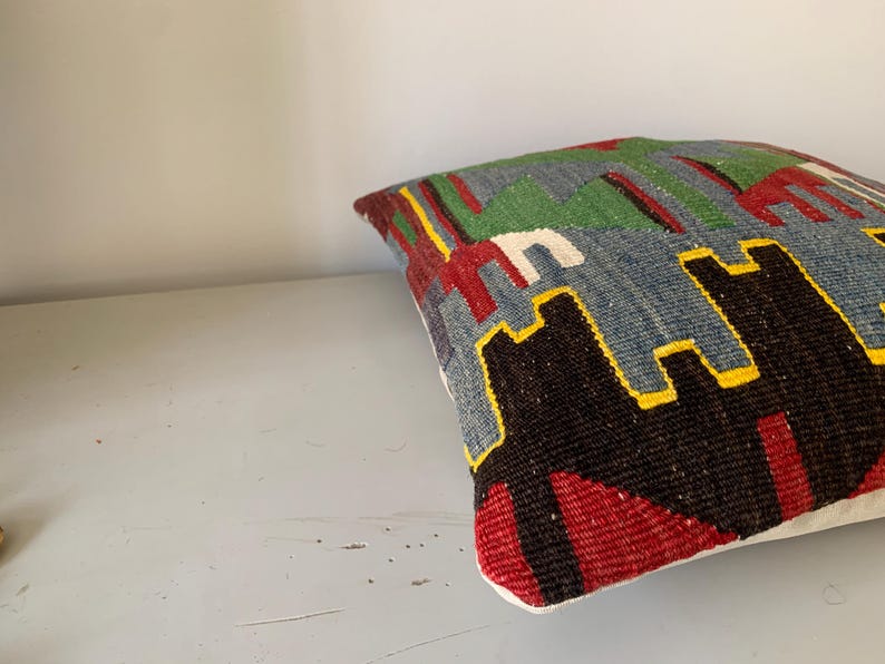 May include: A colorful throw pillow with a geometric pattern in shades of red, green, blue, black, and yellow. The pillow is made of woven fabric and has a white border.