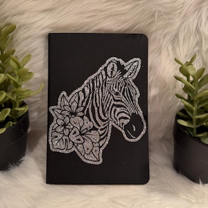 May include: Black notebook with a silver glitter zebra head and floral design. The zebra has black and silver stripes. The notebook is rectangular and has a smooth texture. Two small potted plants flank the notebook.