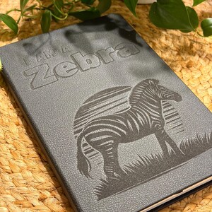 May include: A dark gray journal with the text "I AM A Zebra" embossed on the cover, along with an illustration of a zebra. The zebra is standing in grass. The journal is closed and has a textured surface.