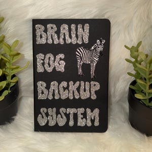 May include: Black notebook with silver glitter text that reads "BRAIN FOG BACKUP SYSTEM". A zebra illustration is on the right side. The notebook is on a white fluffy surface with green plants in black pots on either side.