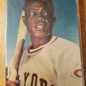 May include: A vintage baseball card featuring a black and white photo of a baseball player wearing a white jersey with the word "YORK" on the front, a black cap with a white "NY" logo, and holding a wooden baseball bat.