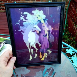 May include: Framed art print featuring a fantasy illustration of a woman with long white hair and a purple cloak, standing beside a white unicorn. The background is a deep purple with blue butterflies. The black frame has a simple design.