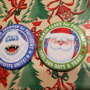 May include: Two round button pins with political campaign slogans. The left pin features a blue background with a white abominable snowman and the text "Abominable Snowman for President, Misfits Unite!" The right pin features a green background with a white Santa Claus and the text "Santa Claus for President, Toys: 365 Days a Year!"
