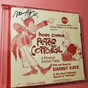 May include: A red and white CD case with a cartoon illustration of Peter Cottontail and a musical Easter fable. The case features the text "Here Comes Peter Cottontail" and "Told and Sung by Danny Kaye".