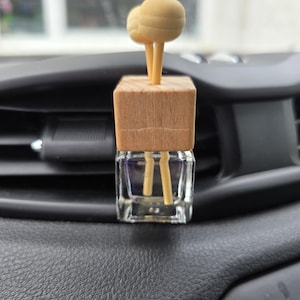 Könnte beinhalten: Ein Auto-Lufterfrischer mit einem Holzsockel in Würfelform und zwei kugelförmigen Holzaufsätzen. Die Basis ist eine kleine, transparente Glasflasche. Der Lufterfrischer steht auf einem schwarzen Armaturenbrett.
