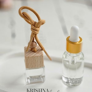 Könnte beinhalten: Eine klare Glasflasche mit goldenem Deckel und einem weißen Tropfer, neben einem quadratischen Holzdiffusor mit einem beigefarbenen Band. Der Diffusor hängt an einem beigefarbenen Band. Die Gegenstände befinden sich auf einer weißen Oberfläche mit dem Text "KRISTINA Korzhenko DESIGN" in Schwarz.