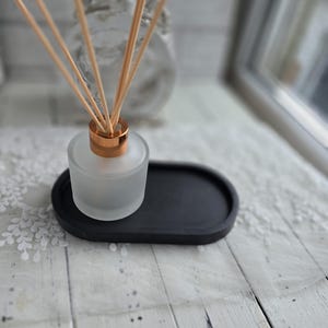 Könnte beinhalten: Ein Diffusor aus mattiertem Glas mit kupferfarbenen Akzenten und Holzstäbchen, auf einem schwarzen ovalen Tablett. Der Diffusor ist für Raumduft und steht auf einer weißen Holzoberfläche.