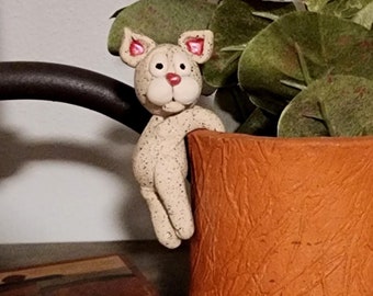 Puede incluir: Una pequeña figurita de cerámica de un gato asomándose desde una maceta de terracota con una planta verde frondosa. La maceta tiene forma de tronco de árbol y tiene una superficie texturizada.