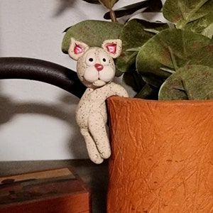 以下が含まれることがあります： 緑の葉のついたテラコッタ製のプランターから顔を出している猫の小さなセラミック製フィギュア。プランターは木の幹の形をしていて、表面はざらざらしている。