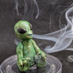 May include: A green alien figurine with large eyes and a wide mouth, sitting in a meditative pose on a clear circular base. Smoke curls up from the base, creating a whimsical and otherworldly scene.
