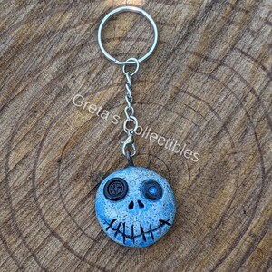 May include: A blue and white clay keychain with a stitched mouth and one black button eye and one blue button eye. The keychain is attached to a silver key ring.