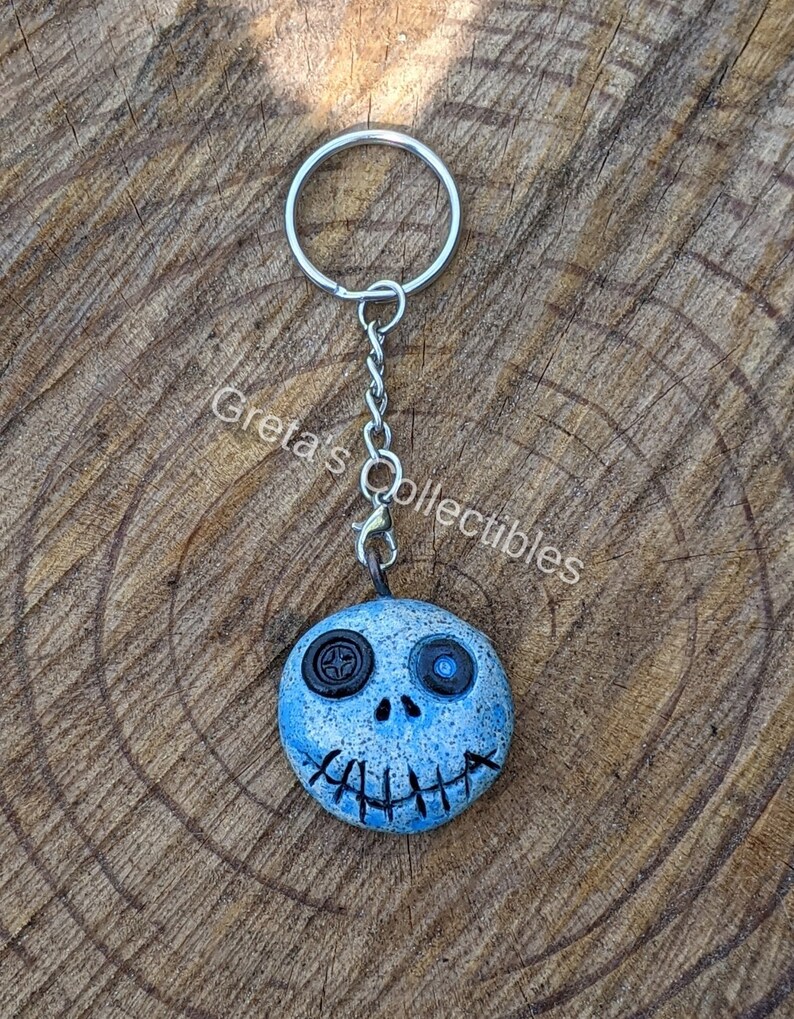 May include: A blue and white clay keychain with a stitched mouth and one black button eye and one blue button eye. The keychain is attached to a silver key ring.