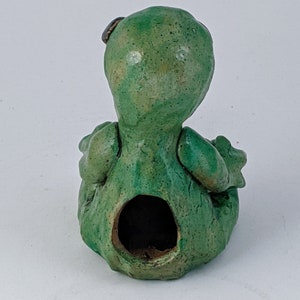 May include: Green ceramic figurine of a sitting creature with a hole in the back. The creature has a textured surface and a small brown detail on its head.