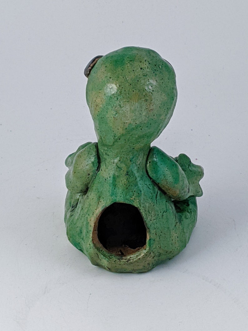 May include: Green ceramic figurine of a sitting creature with a hole in the back. The creature has a textured surface and a small brown detail on its head.