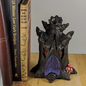 May include: A black ceramic tree house with a purple door and red mushroom accents. The tree house is sitting on a wooden surface next to two books. The book on the left is titled "15 Minute Low Carb Recipes Dana Carpender". The book on the right is titled "When We Were Very Young Milne Dutton".