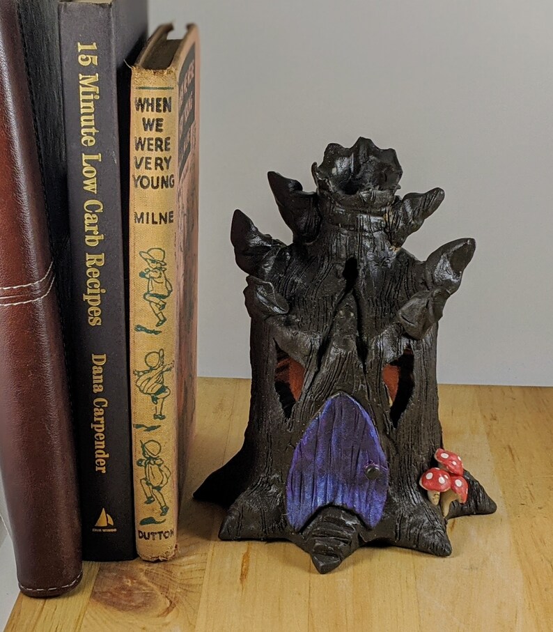 May include: A black ceramic tree house with a purple door and red mushroom accents. The tree house is sitting on a wooden surface next to two books. The book on the left is titled "15 Minute Low Carb Recipes Dana Carpender". The book on the right is titled "When We Were Very Young Milne Dutton".