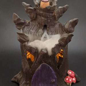 May include: A ceramic incense burner shaped like a tree stump with a purple door and a small red mushroom. Smoke is rising from the top of the burner.